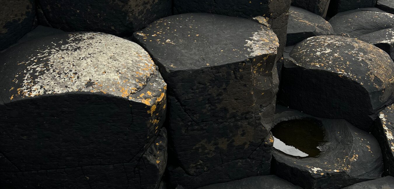 Large, dark hexagonal basalt columns with rough surfaces, some covered in pale lichen, form the foundations of a growth journey. A small pool rests in a natural depression; soft lighting highlights the rocks’ textures.