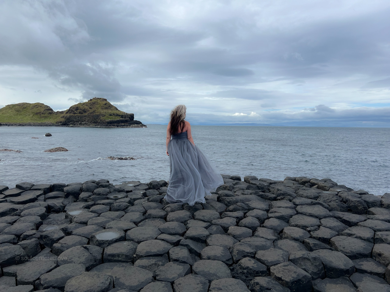 A woman in a flowing gray dress stands on large, hexagonal basalt rocks by the sea, embodying a Soul-Centered Freedom Journey as she gazes toward the ocean under a cloudy sky, with a grassy hill in the distance.